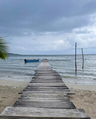 Fototapeta premium A weathered wooden pier stretches in the calm Caribbean Sea at Bocas del Toro, Panama. A small blue boat floats, cloudy sky.