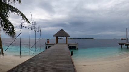 Luxury resort photo of Maldives tropical paradise island in Indian Ocean with hotel territory, palm trees  on beach, wooden bridge. Beautiful photography, azure ocean, sky with clouds.