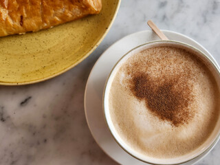 overhead close-up shot of a cappuccino with cinnamon on top, next to a pastry on a yellow plate.