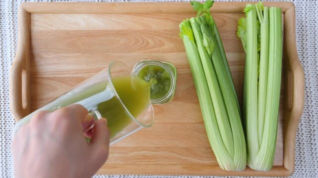 Woman's hand pouring freshly squeezed green celery juice from a glass pitcher into a jar on a wooden serving tray, a healthy lifestyle and detox diet concept for natural weight loss