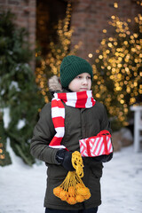 Happy cute teenager boy with mesh bag of tangerines standing near by spruce Christmas tree. Holiday celebrate concept.