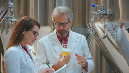 Two scientists in lab coats inspecting the quality of a beer sample, one man holding a beaker of beer and a woman taking notes, working on brewing process control