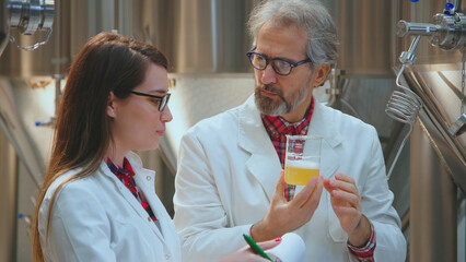 Two scientists in lab coats inspecting a beer sample in a beaker, analyzing its quality and characteristics in a modern brewery laboratory, performing scientific research