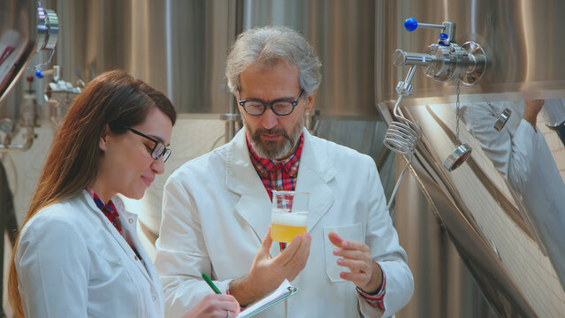 Two scientists in lab coats inspecting the quality of a beer sample, one man holding a beaker of beer and a woman taking notes, working on brewing process control