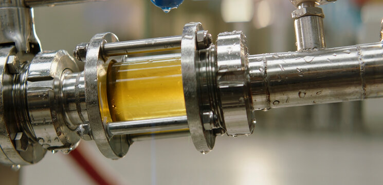 Brewer's wort or beer flowing through a clear sight glass in a stainless steel pipe at a modern brewery, depicting industrial production and manufacturing of alcoholic beverages