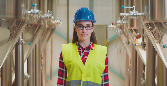 Young woman engineer wearing safety hardhat and high-visibility vest standing in a modern factory setting with large stainless steel tanks in the background, looking at camera