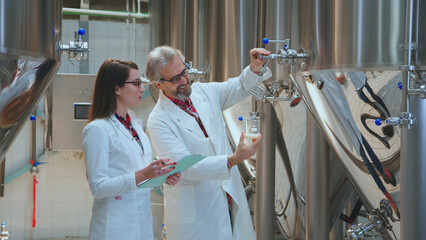 Brewery workers in lab coats performing quality control on a beer sample drawn from large fermentation tanks, ensuring product consistency and adherence to brewing standards