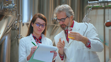 Two scientists in lab coats inspect beer quality in a modern brewery, taking notes on a clipboard while monitoring fermentation tanks and conducting quality control tests