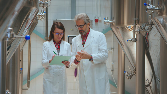 Brewery quality control team members wearing lab coats, collaborating and inspecting samples in a craft beer production facility with large stainless steel tanks