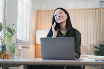 Young woman smiling while talking on phone at laptop in bright home office, relaxed and productive