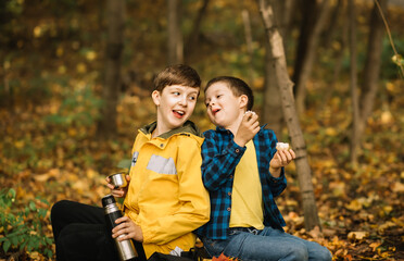 Little kid boys hiking in the forest relax sitting on stump and drinks from thermos in autumn forest