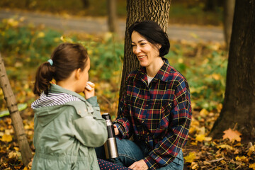 Mother with kid child girl hiking in autumn forest relax sitting on stump, drinks from thermos and eating sandwich in autumn forest.