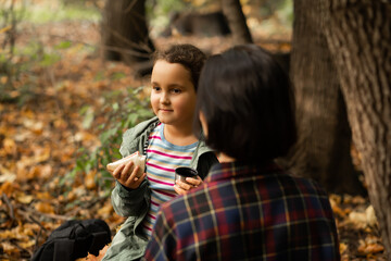 Mother with kid child girl hiking in autumn forest relax sitting on stump, drinks from thermos and eating sandwich in autumn forest.