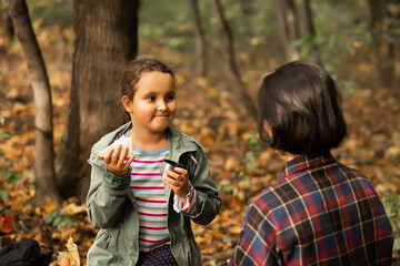 Mother with kid child girl hiking in autumn forest relax sitting on stump, drinks from thermos and eating sandwich in autumn forest.