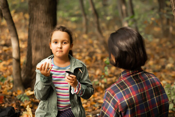 Mother with kid child girl hiking in autumn forest relax sitting on stump, drinks from thermos and eating sandwich in autumn forest.