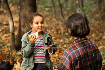 Mother with kid child girl hiking in autumn forest relax sitting on stump, drinks from thermos and eating sandwich in autumn forest.