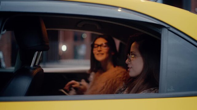 Two stylish young women getting into the back seat of a yellow taxi cab at night and driving away