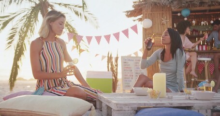 Two women sipping beer and lime cocktail in beach bar lounge, with pallet table and cushions