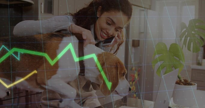 Woman balancing beagle dog and analyzing stock graphs on laptop in home kitchen, with smartphone