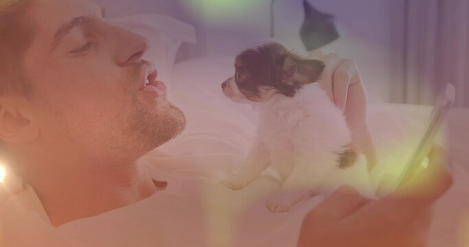 Man balancing small puppy on bed while holding smartphone in bedroom with pillows, lamp, copy space - Powered by Adobe