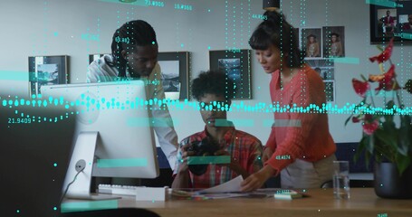 Leaning coworkers reviewing prints and camera at studio table, with monitor and potted plant