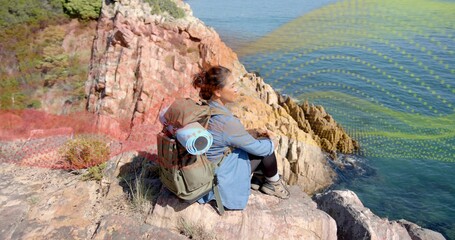 Sitting hiker wearing light denim jacket with olive green backpack on rocky cliff overlooking sea