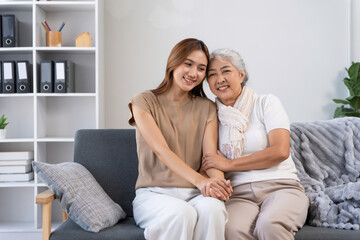 Happy Asian mother and adult daughter smiling, holding hands, and embracing on a sofa, showing love and care