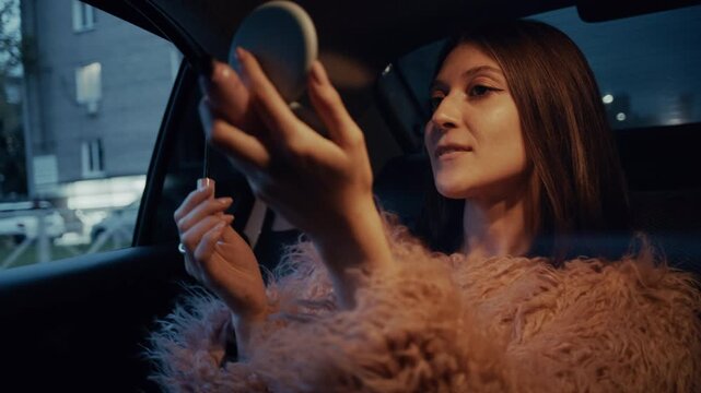 Young woman applying lipstick and mascara in the back of a moving car in the evening