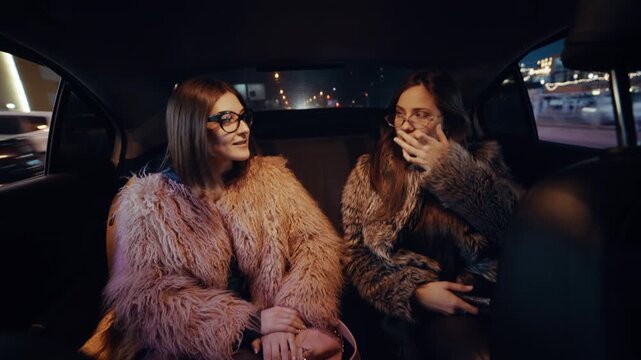 Two stylish girls in luxurious fur coats sit in the back seat of a taxi, laughing and chatting as the bright city lights flash through the windows at night.
