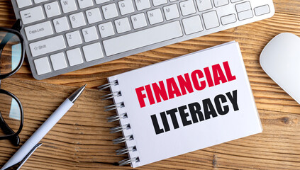 FINANCIAL LITERACY text on spiral notepad with keyboard, mouse, glasses and pen on wooden desk
