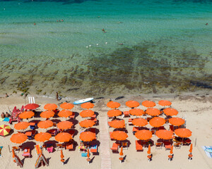 Aerial view of a beach club with orange umbrellas and orange sunbeds in Italy. There are many people on holiday on this beautiful sunny summer day. The Mediterranean Sea is beautiful and clear.