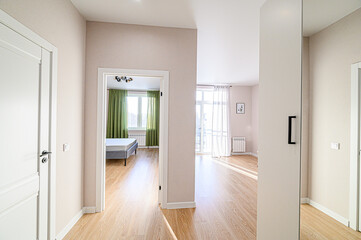 A bright, modern apartment interior with light wood floors. Seen through doorways are a bedroom with green curtains and a living room with sheer white curtains