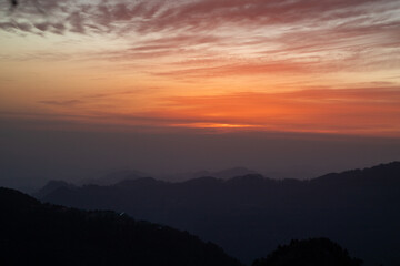 A breathtaking mountain sunset with layers of dark hills fading into the horizon under a dramatic orange and red sky. The soft glow of the setting sun and the textured clouds create a peaceful and ser