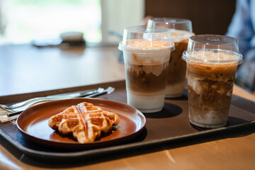 Close up and selective focus glasses of iced coffee with croffle in plate on tray in coffee shop.