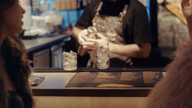 Male bartender preparing alcoholic drinks for two women sitting at the counter in a modern bar
