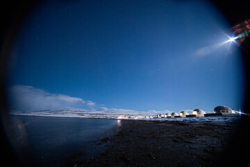 A row of illuminated glamping domes along a snowy, rocky shoreline under a dark, starry night sky with a bright lens flare