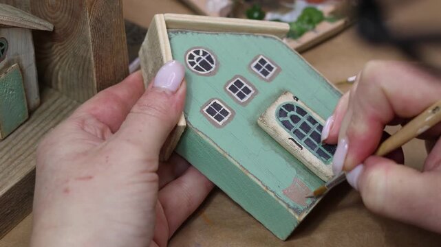 Close up of woman's hands artfully painting small wooden toy house with brush, demonstrating arts and crafts skills to create a decorative home decor, showcasing creativity and hobby