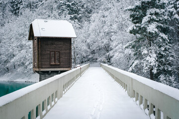 Snowy bridge leads to a wooden house by a lake.