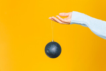 Woman's hand holding a shiny blue Christmas ornament against a bright yellow background.