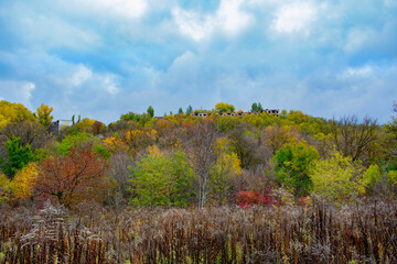 Fototapeta premium A picturesque view of a forest with trees in vibrant autumn colours and an old abandoned house visible in the distance on a hillside under a cloudy blue sky.