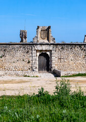 The medieval stone wall and entrance of the weathered Castillo de los Condes in Chinchón, Spain, framed by a lively field of yellow wildflowers and surrounded by a dirt road