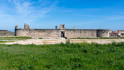 The medieval stone wall and entrance of the weathered Castillo de los Condes in Chinchón, Spain, framed by a lively field of yellow wildflowers and surrounded by a dirt road