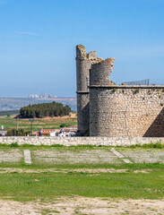 The medieval stone wall and entrance of the weathered Castillo de los Condes in Chinchón, Spain, framed by a lively field of yellow wildflowers and surrounded by a dirt road