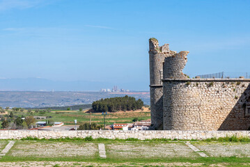 The medieval stone wall and entrance of the weathered Castillo de los Condes in Chinchón, Spain, framed by a lively field of yellow wildflowers and surrounded by a dirt road
