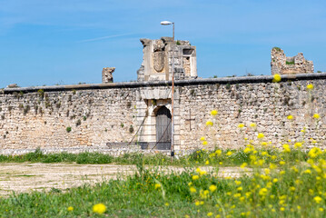 The medieval stone wall and entrance of the weathered Castillo de los Condes in Chinchón, Spain, framed by a lively field of yellow wildflowers and surrounded by a dirt road