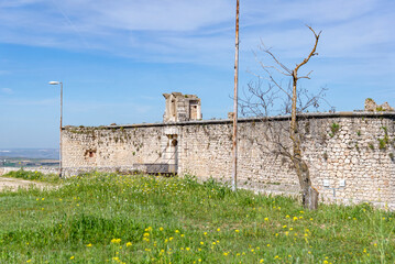 The medieval stone wall and entrance of the weathered Castillo de los Condes in Chinchón, Spain, framed by a lively field of yellow wildflowers and surrounded by a dirt road