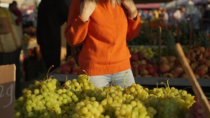 Sunny autumn day at local street market with woman in an orange sweater choose fresh, ripe green grapes from vendor's stall full of delicious and healthy organic produce - Powered by Adobe