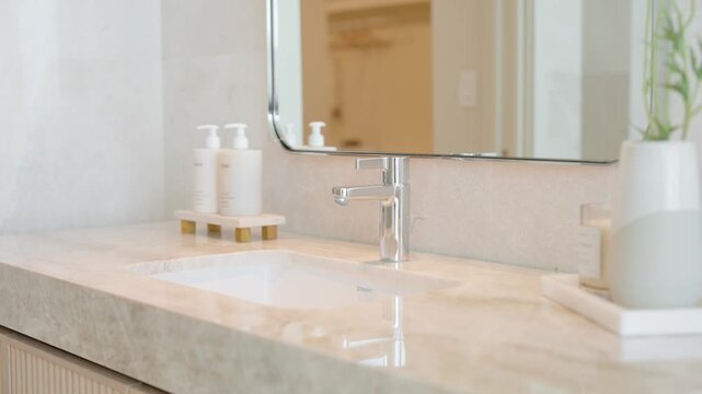 Clean and modern bathroom countertop with light stone surface and central chrome faucet. Above it hangs rectangular mirror with thin frame. Two white pump bottles