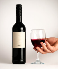 Hand holds glass of red wine beside dark bottle on white background, symbolizing elegance, taste, and refined lifestyle.
