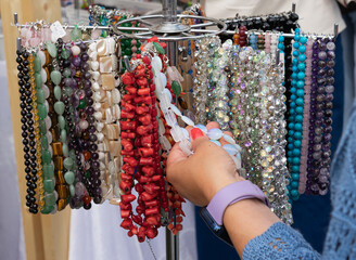 Collection of colorful gemstone necklaces and bracelets on jewelry rack, close-up of woman’s hand choosing crystal accessory at market